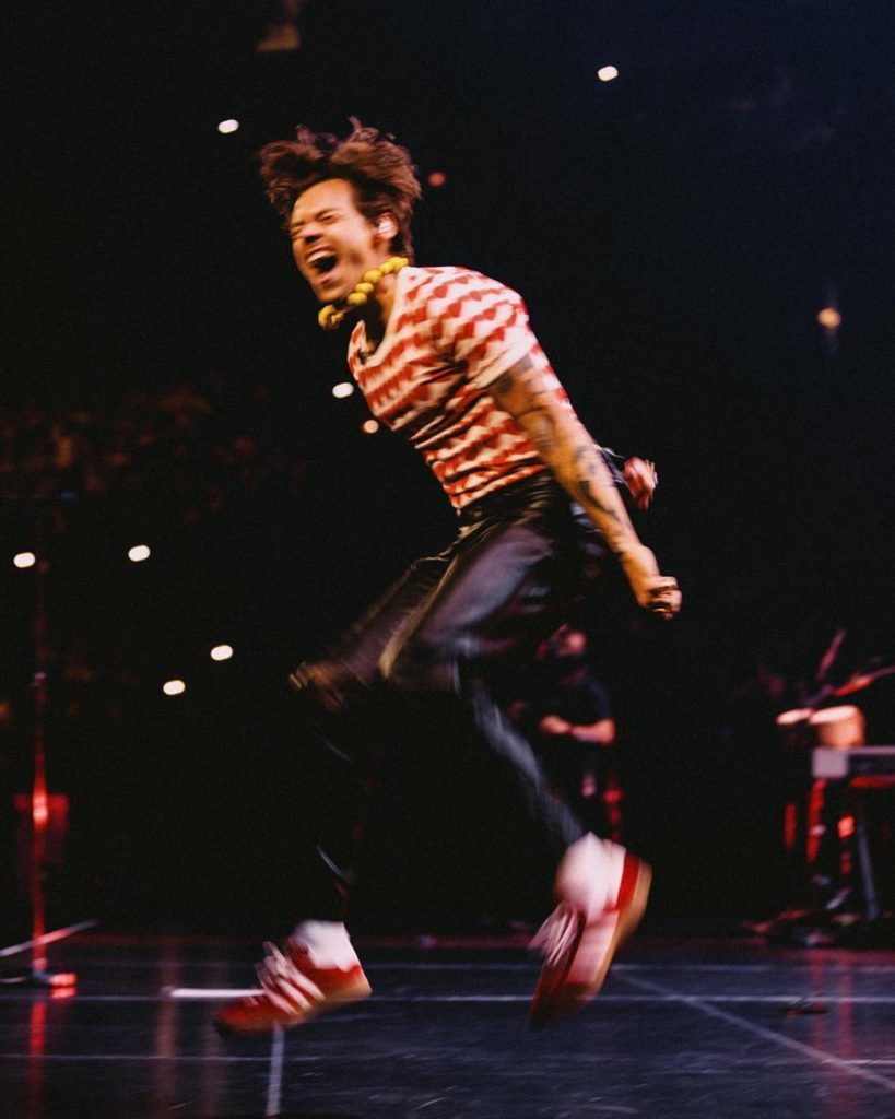 O cantor Harry Styles, um homem jovem, branco, com cabelo liso castanho, está cantando e dançando durante um show.  Ele usa uma camiseta listrada vermelha e branca, calça preta e um tênis vermelho da Adidas.  - Metrópoles